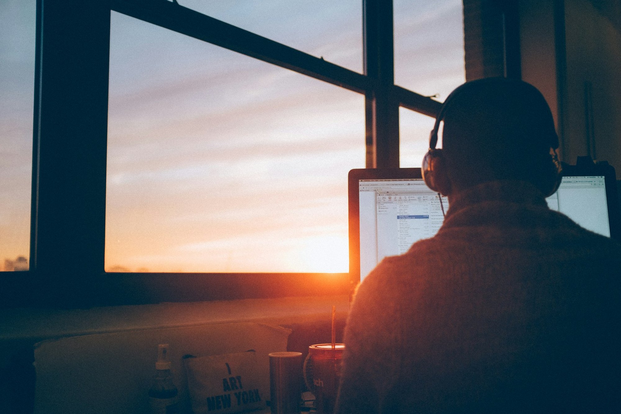 Person sitter framför en laptop med hörlurar vid ett fönster i soluppgången