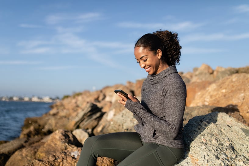 Kvinna på sten vid havet med mobil i handen